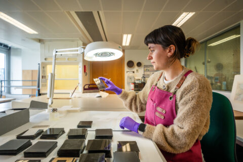 A conservator sits at a bench inspecting a glass plate negative. Further glass plate negatives are neatly arranged in piles next to her.
