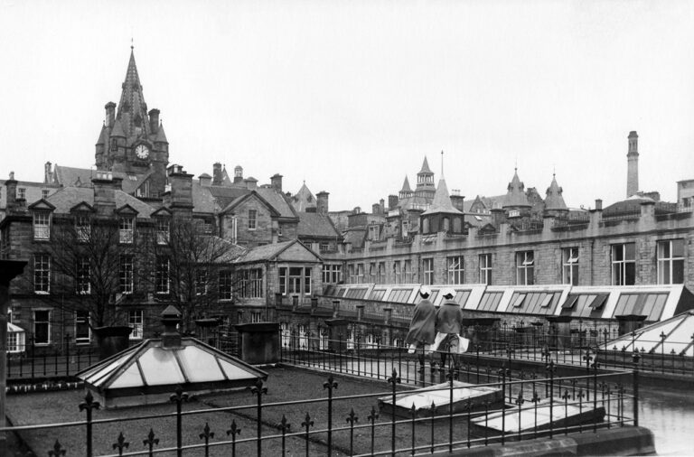 black and white photograph of two nurses walking across a rooftop walkway