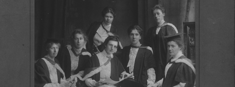 A black and white photograph of 7 women in assorted white blouses and graduation gowns. Two are wearing graduation caps. One in the foreground holds a degree certificate.